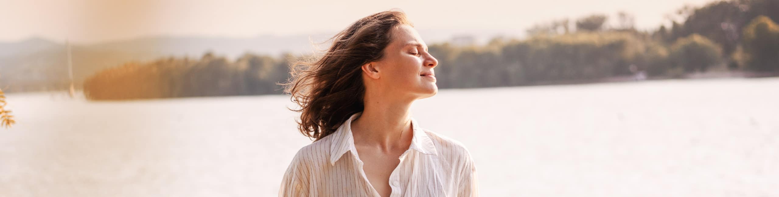 Eine Frau steht im Freien an einem See und genießt die Sonne mit geschlossenen Augen.