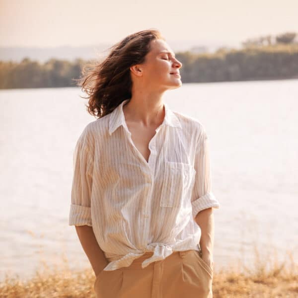 Eine Frau steht im Freien an einem See und genießt die Sonne mit geschlossenen Augen.