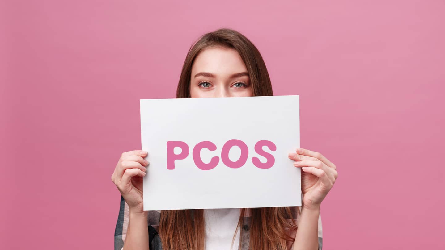 Frau hält das Schild „PCOS“ vor rosa Hintergrund